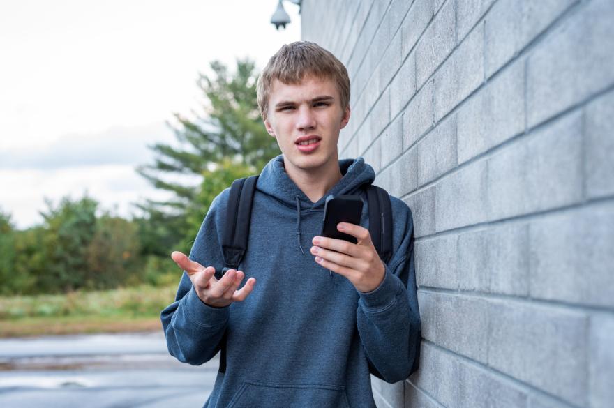 Jongen leunend tegen muur met gsm in hand, vragend gebaar.